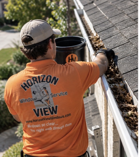 Professional gutter technician in bright orange polo and khaki hat clearing leaves from roofline gutters on a Dallas Fort Worth brick home
